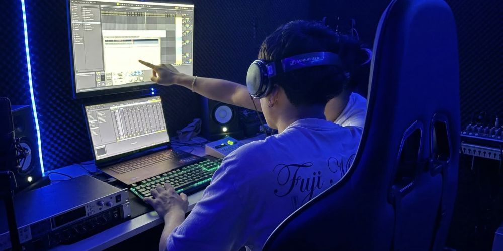 Un ingeniero de audio imparte una formación a un alumno utilizando auriculares Sennheiser, monitores duales, Ableton Live, un teclado mecánico RGB y tratamiento acústico.