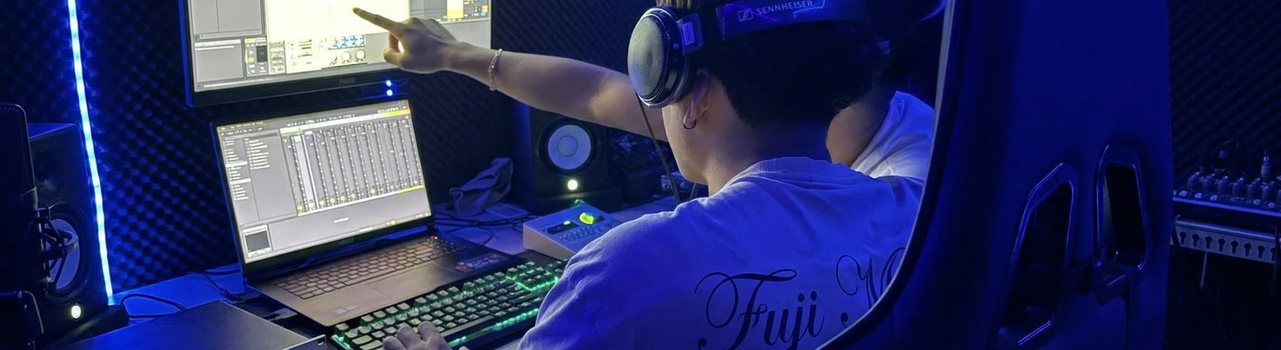 Un ingeniero de audio imparte una formación a un alumno utilizando auriculares Sennheiser, monitores duales, Ableton Live, un teclado mecánico RGB y tratamiento acústico.