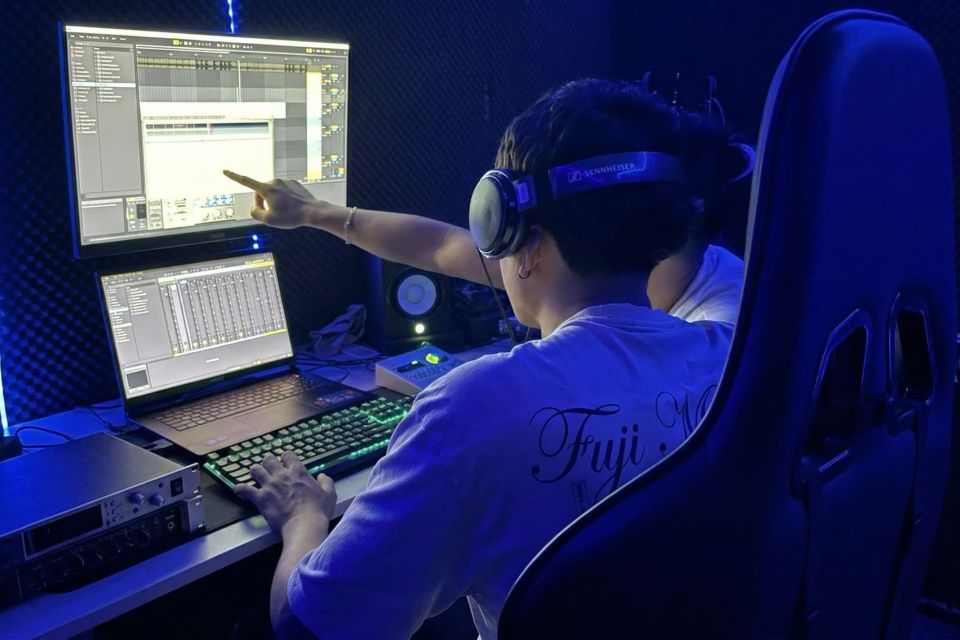 Un ingeniero de audio imparte una formación a un alumno utilizando auriculares Sennheiser, monitores duales, Ableton Live, un teclado mecánico RGB y tratamiento acústico.